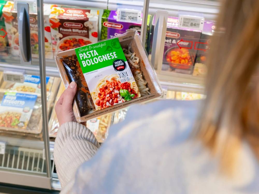 En person holder en ferdig pakke med pasta Bolognese i en matbutikk, med frysedisk og andre frosne matvarer i bakgrunnen.