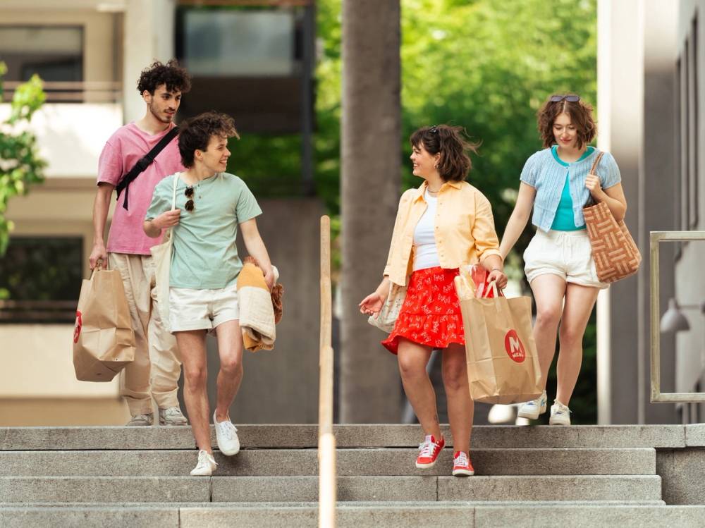 Fire unge voksne går nedover trappen utendørs med handleposer og smiler, omgitt av grønne trær og bygninger.