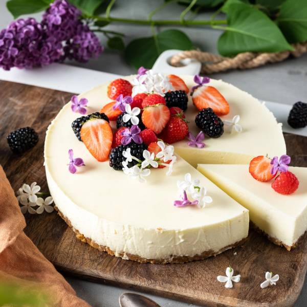 En ostekake pyntet med friske bær og små spiselige blomster på en rund treplate.