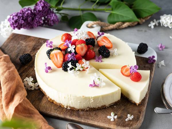 En ostekake pyntet med friske bær og små spiselige blomster på en rund treplate.