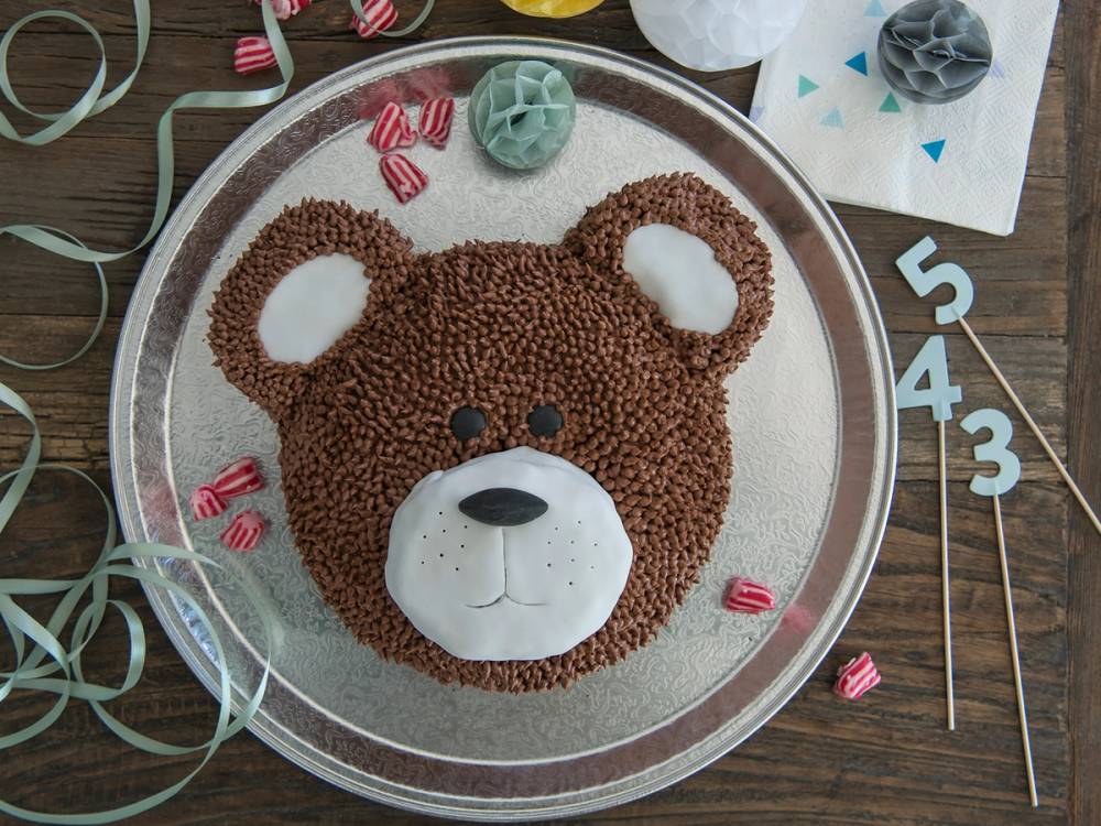 En kake formet som en bjørn med brune og hvite detaljer, dekorert med små godterier og festpynt rundt på et bord.
