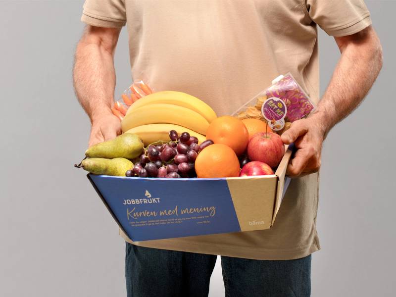 En person holder en eske med ulike frukter som bananer, druer, epler, og appelsiner.