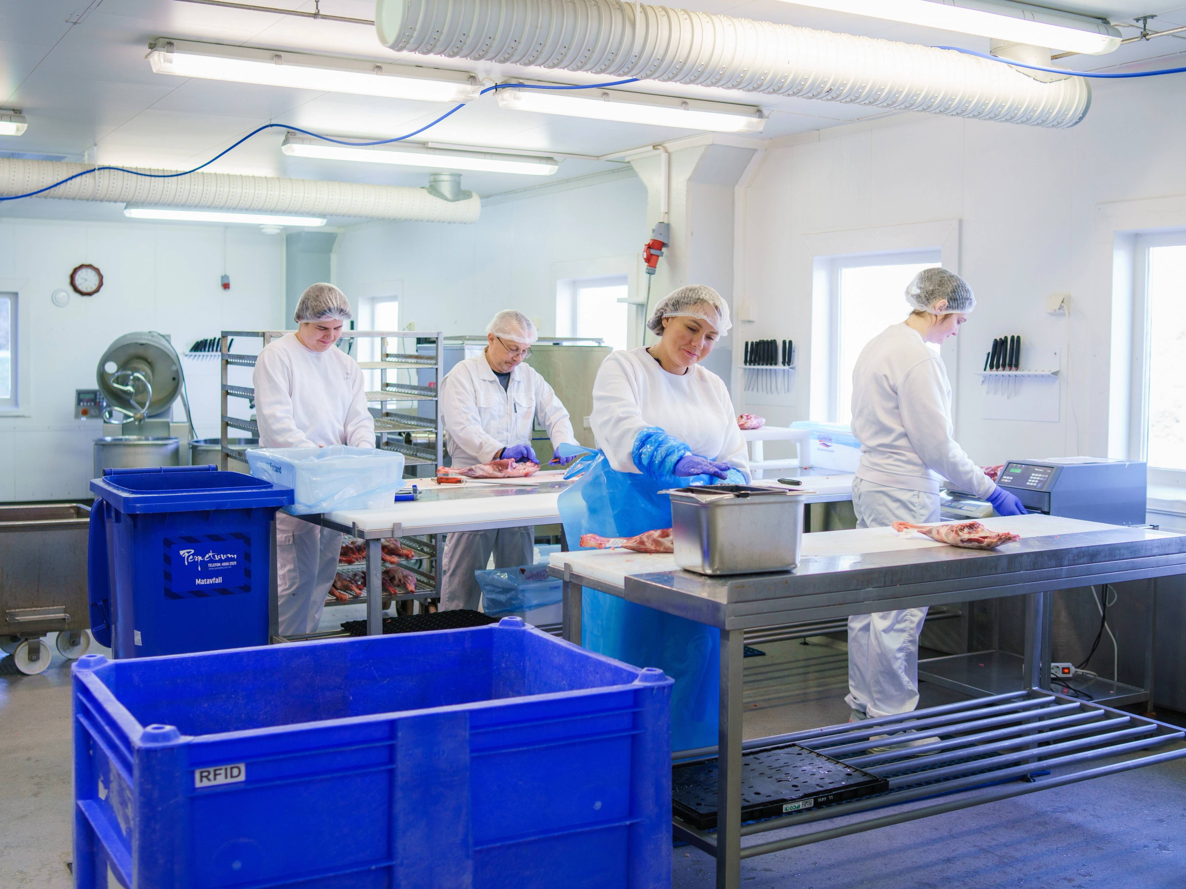 Personer i hvite kjeledresser jobber i et industrielt kjøkken eller laboratorium med arbeidsbord og utstyr.