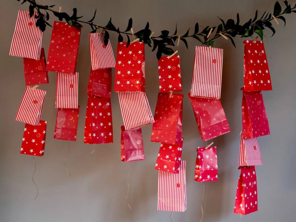 Hengende små gaveposer og kort i røde og hvite mønstre, dekorert med grønt bladbånd over en beige vegg.