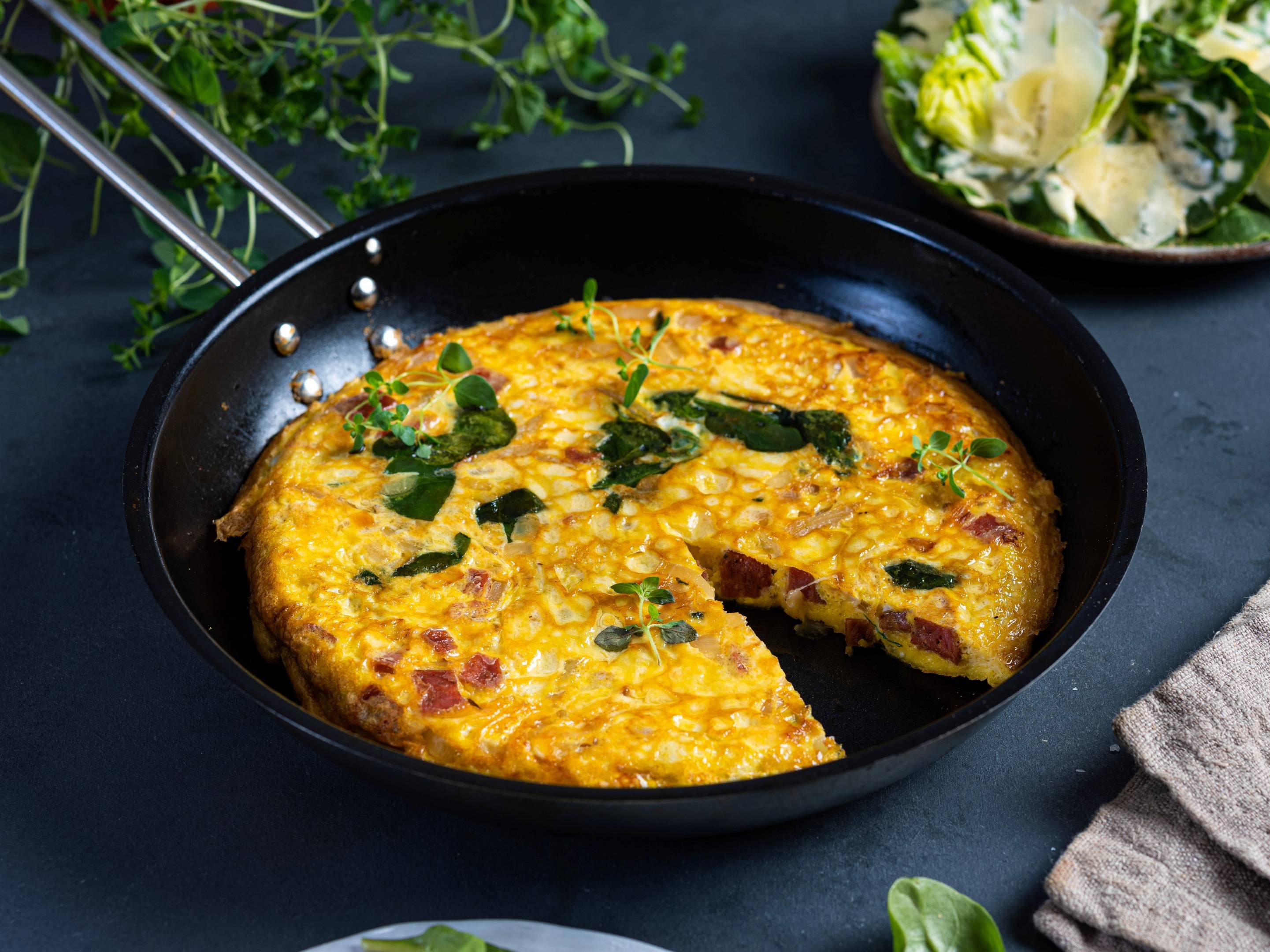 En gyllenbrun omelett med grønne urter i en svart stekepanne, med en tallerken med hvite blomster i bakgrunnen.