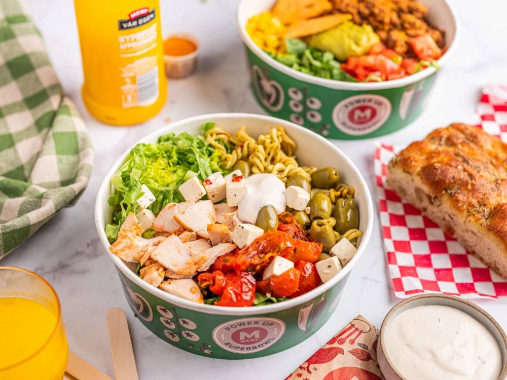 En salat med kylling, grønnsaker og oliven i en grønn salatbolle, med en pizza og en flaske med dressing i bakgrunnen.