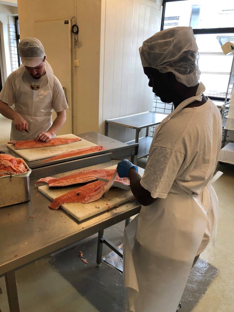 Personal skjærer laks i et kjøkkenmiljø med to personer iført hvite kledninger og hårnetter.