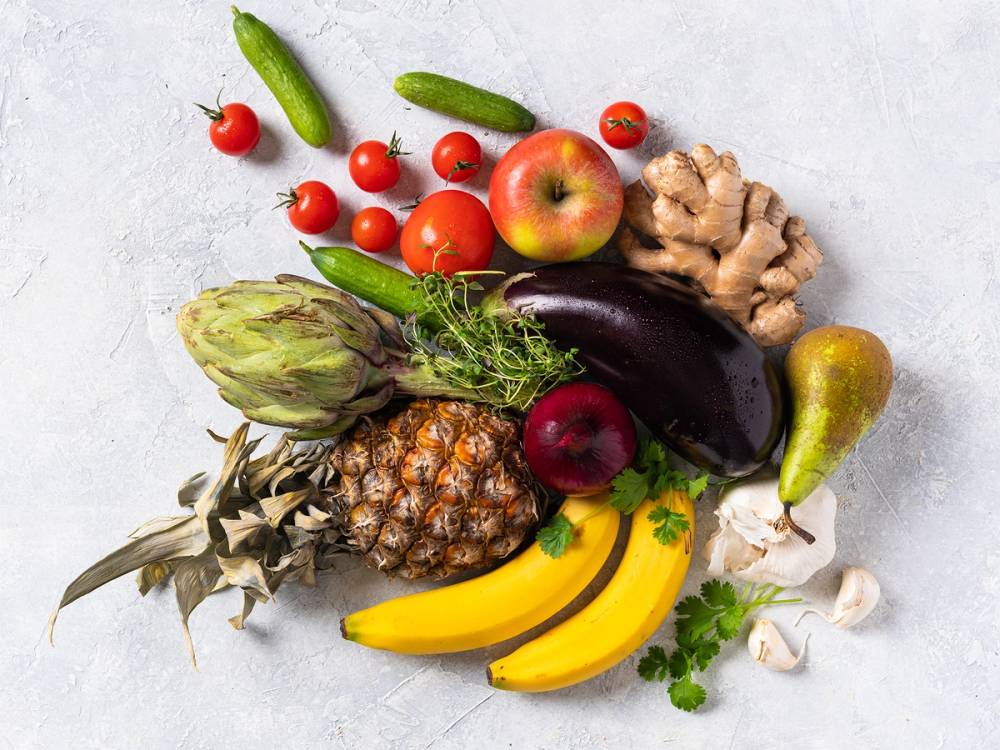 Ferske frukt og grønnsaker som tomater, agurk, epler, ananas, bananer, hvitløk, ingefær, sopp og persille lagt ut på en lys bakgrunn.