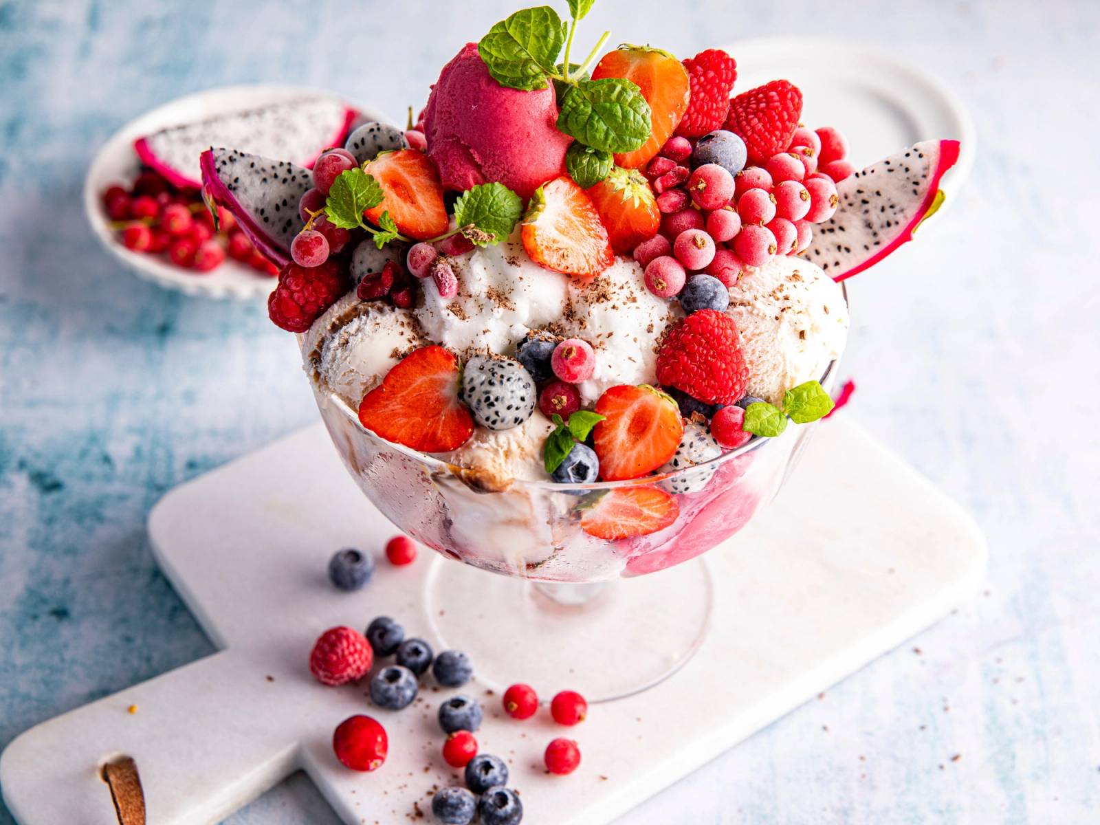 En stor skål med iskrem toppet med ulike frukter som jordbær, bringebær, blåbær og kiwi, med flere bær rundt på en hvit overflate.