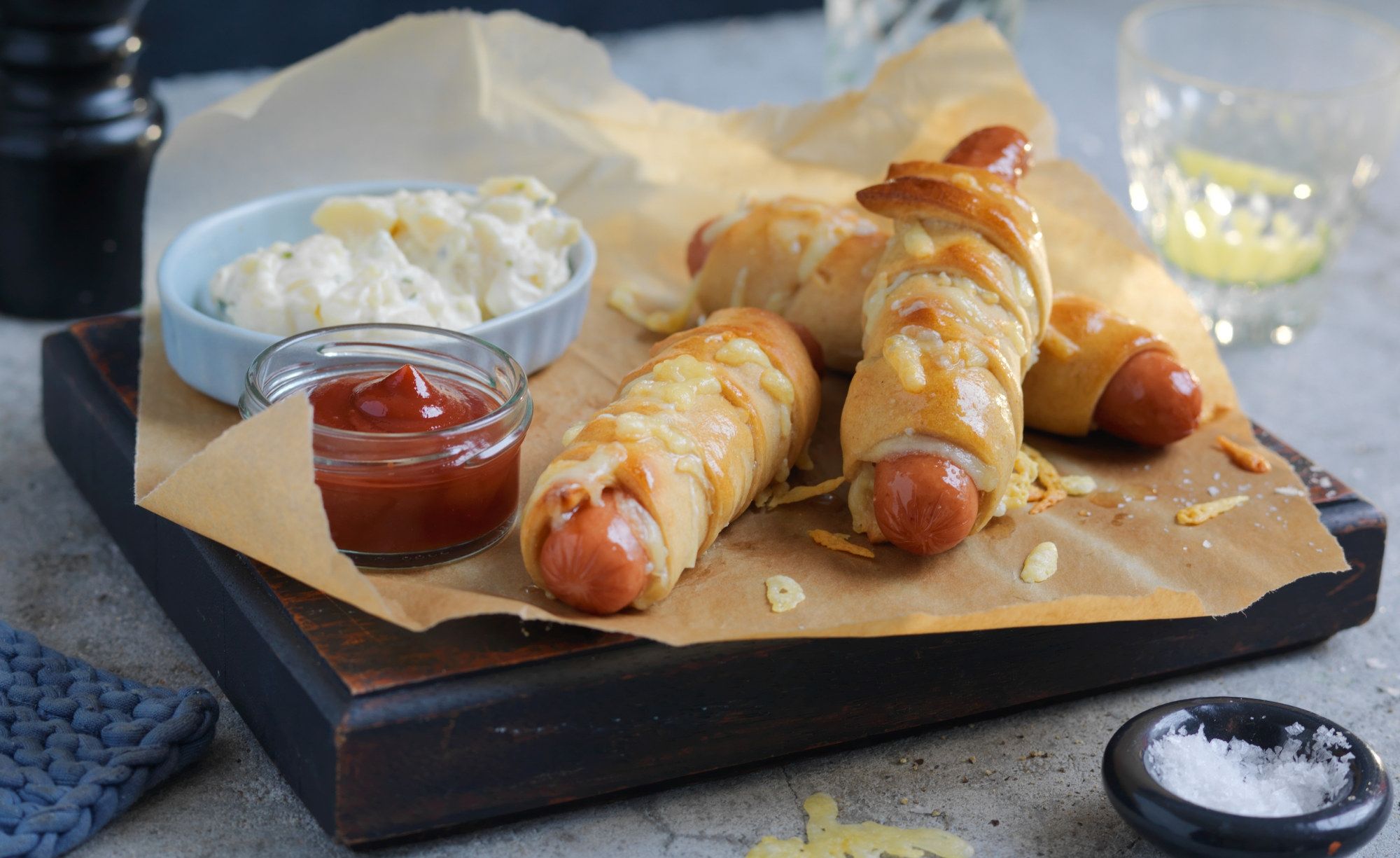 Innbakte pølser med revet ost på en serveringsplate med tilbehør som ketchup og potetsalat.