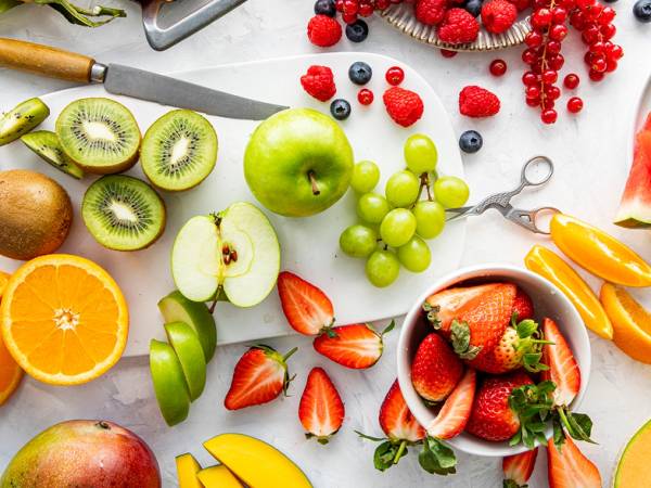 Fargerik samling av frukt og grønt, inkludert vannmelon, kiwi, eple, druer, jordbær, appelsin, og sitron, på et hvitt bord.