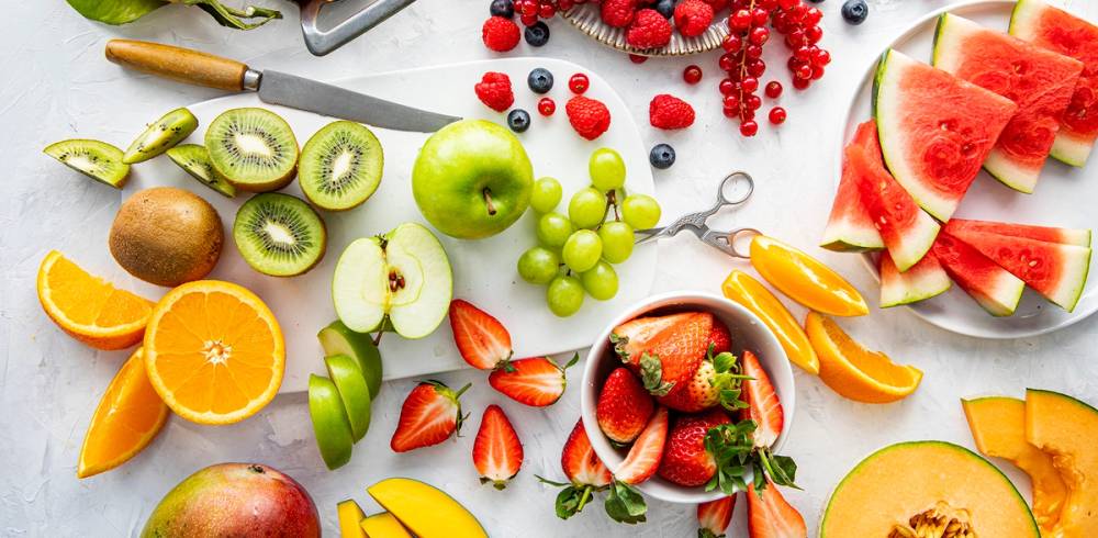 Fargerik samling av frukt og grønt, inkludert vannmelon, kiwi, eple, druer, jordbær, appelsin, og sitron, på et hvitt bord.