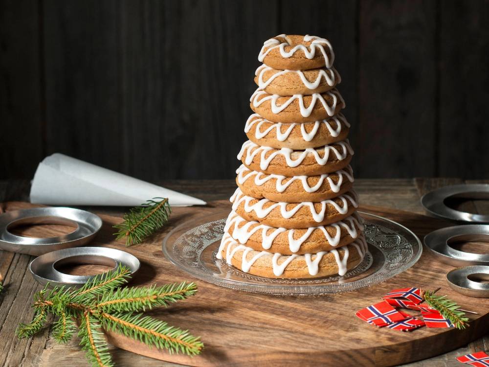 En kransekake dekorert med hvit glasur står på et rundt bord med julepynt og små kakeformer rundt.