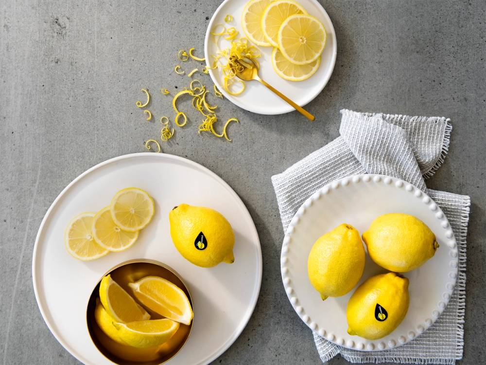 Sitronskiver og en hel sitron på hvit tallerken, med en skje med sitronskall og sitronzest på en liten tallerken, på et grått bord med et kjøkkenhåndkle ved siden av.