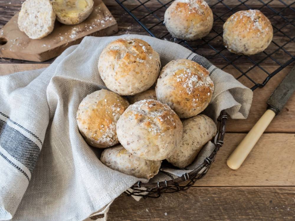 Rundstykker med melis på toppen ligger i en kurv på et bord med et kjøkkenhåndkle og et knivhåndtak i nærheten.
