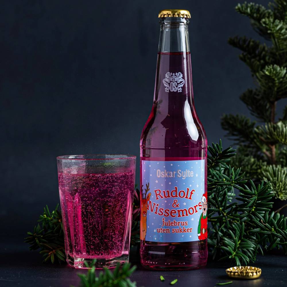 En flaske med julebrus og et glass fylt med rosa julebrus, omgitt av julegrener og en liten julegave, på en mørk bakgrunn.