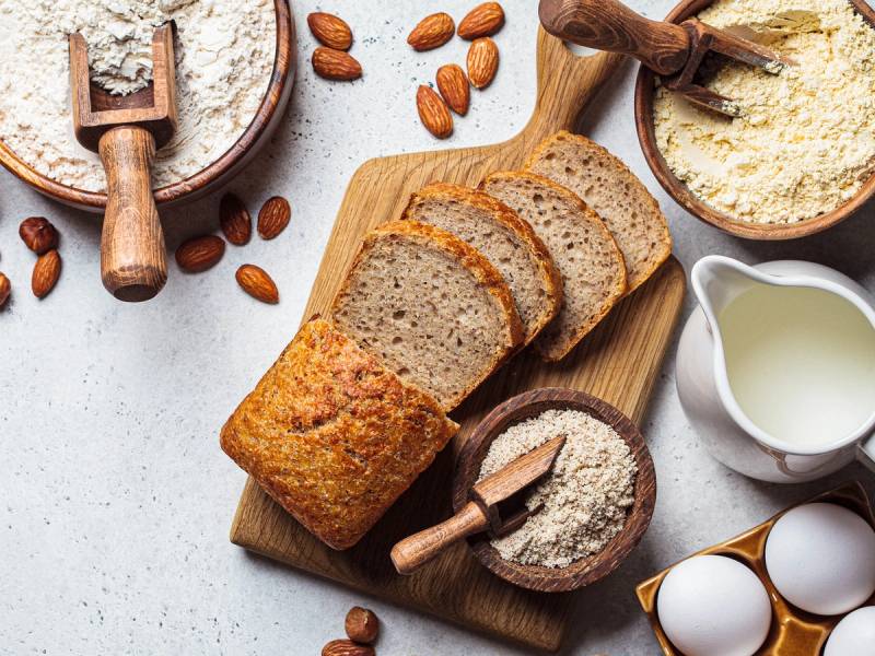 Glutenfritt brød og mel på et kjøkkenbord med nøtter og egg rundt.