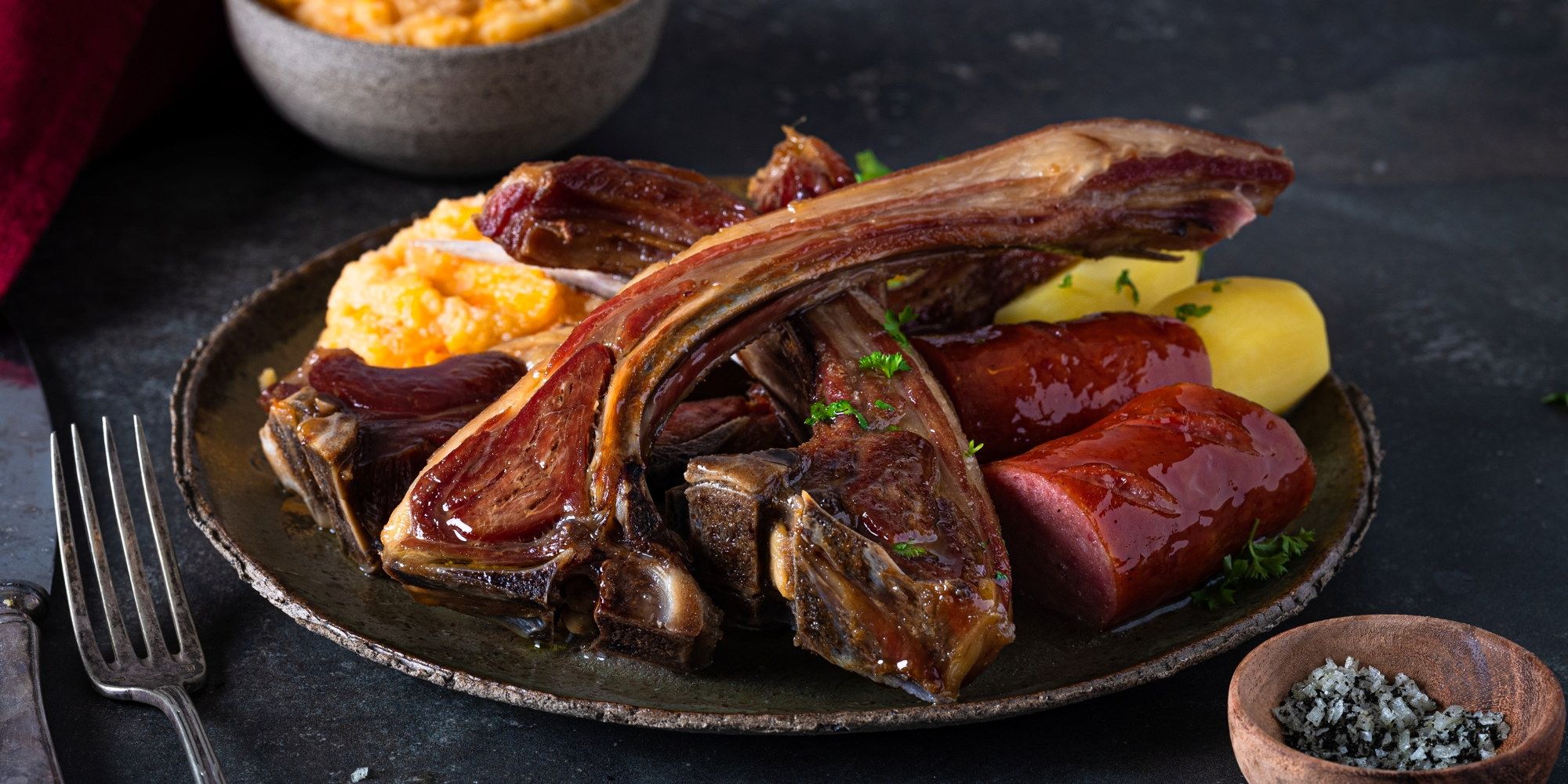 Pinnekjøtt med kokte poteter og gulrot på en svart tallerken, servert med litt grønt som pynt.