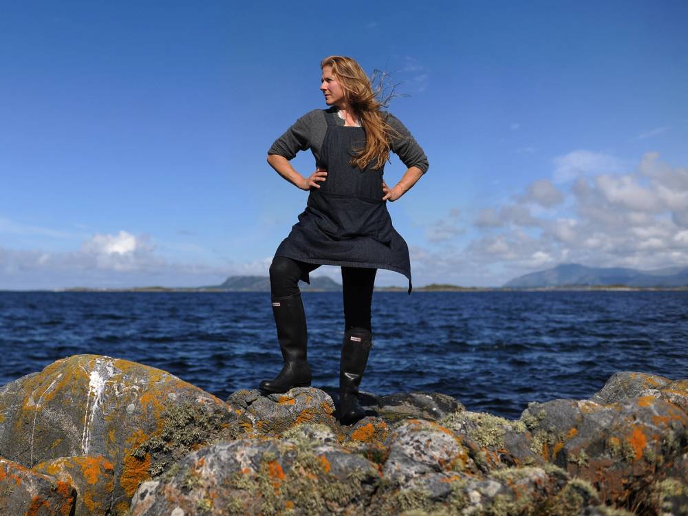 En person står på store steiner ved vannet i Løvika, med armene på hoftene og utsikt mot sjøen og himmelen.