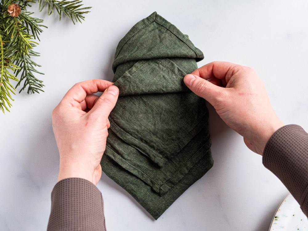 Hender som folder en grønn serviett i form av et juletre på et hvitt bord med noen grønne grener i hjørnet.