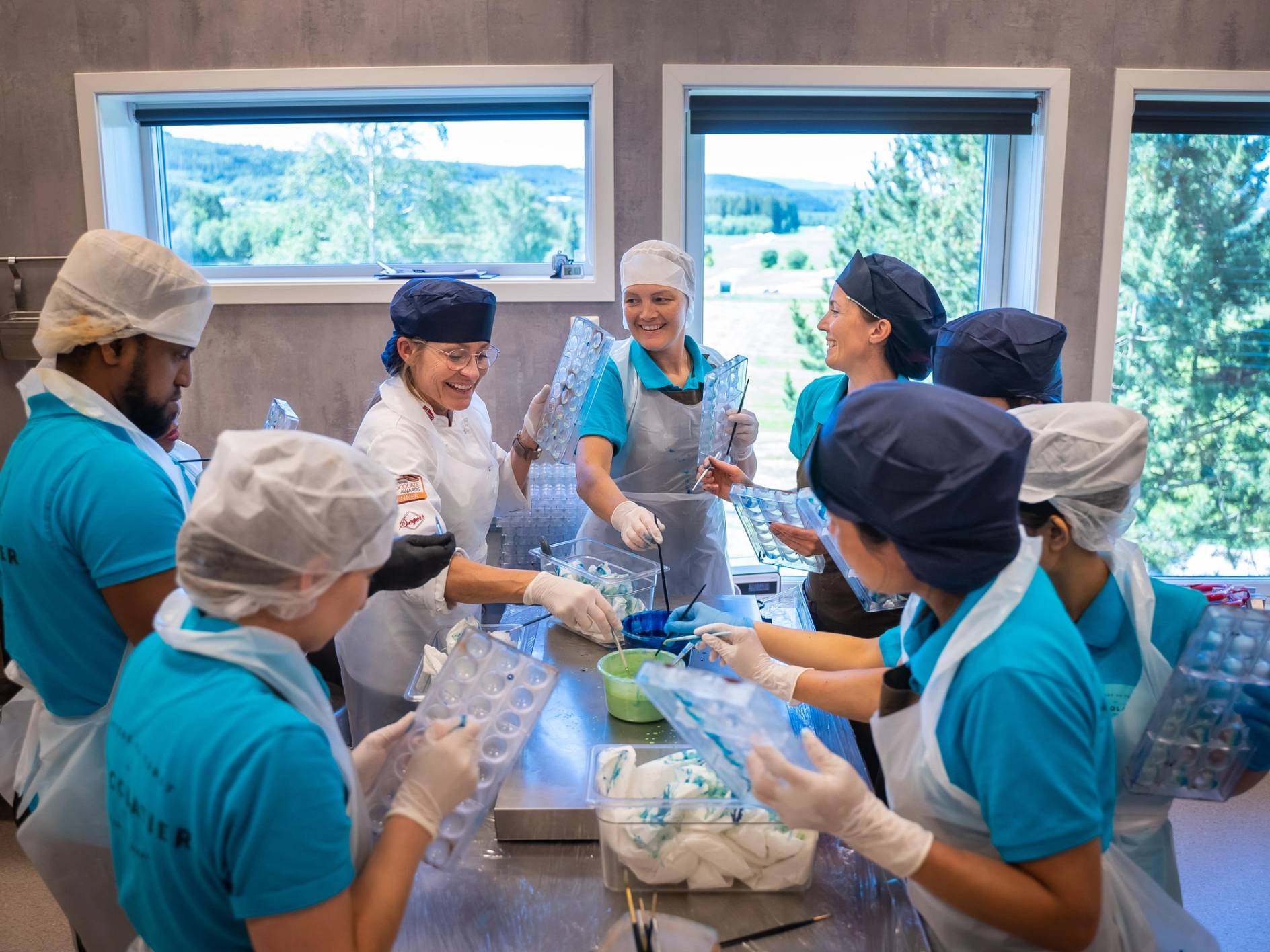 En gruppe jenter i blå og hvite kokkeklær som lager mat sammen i et kjøkken, med store vinduer i bakgrunnen som viser grønt landskap.