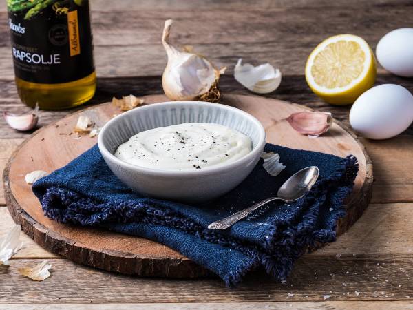 En skål med aioli på et trebrett, omgitt av hvitløk, sitron, egg og en flaske med aiolisaus, på et bord med en blå duk.