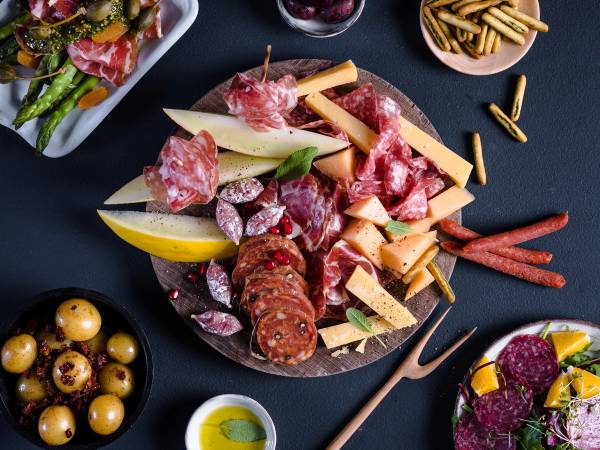 Et fat med ulike typer spekemat, ost og kjeks, omgitt av små skåler med oliven og annet tilbehør, på et svart bord.