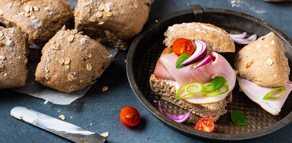 Lunsjbiter med skinke, grønnsaker og ost på en tallerken, omgitt av grove brødskiver og cherrytomater