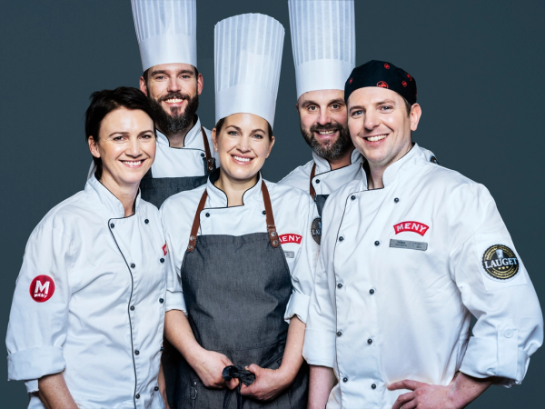 Fire personer i kokkeuniformer står sammen, og en av dem har en grå forkle, mot en mørk bakgrunn.