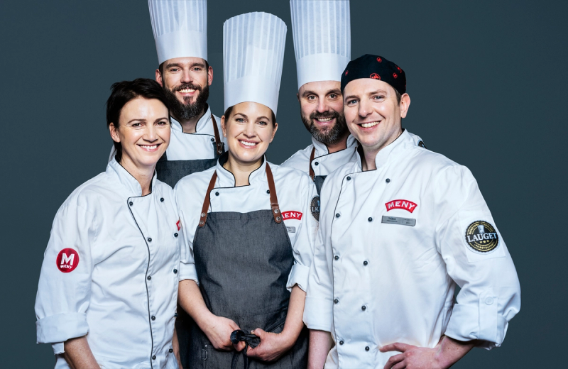 Fire personer i kokkeuniformer står sammen, og en av dem har en grå forkle, mot en mørk bakgrunn.