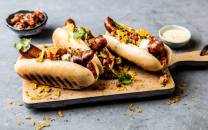 Tre cheesymexican hot dogs med tilbehør på en svart skjærefjøl, med en liten skål med salsa i bakgrunnen.