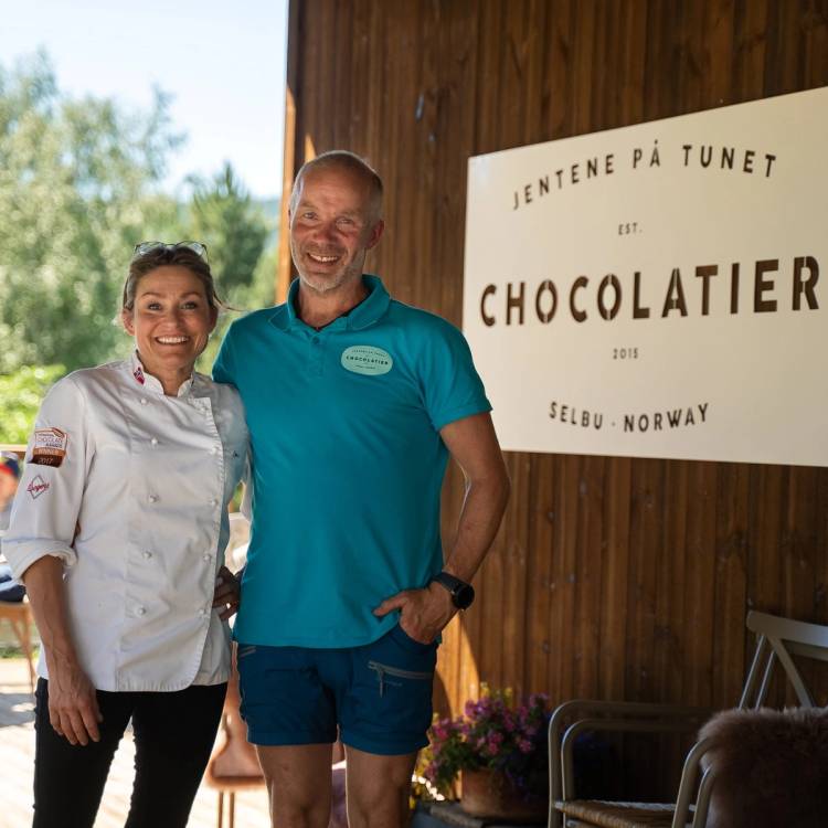 To personer står ved en tavle som sier 'Jentene på Tunet' og 'Chocolatier' utenfor en bygning, med grønne trær i bakgrunnen.