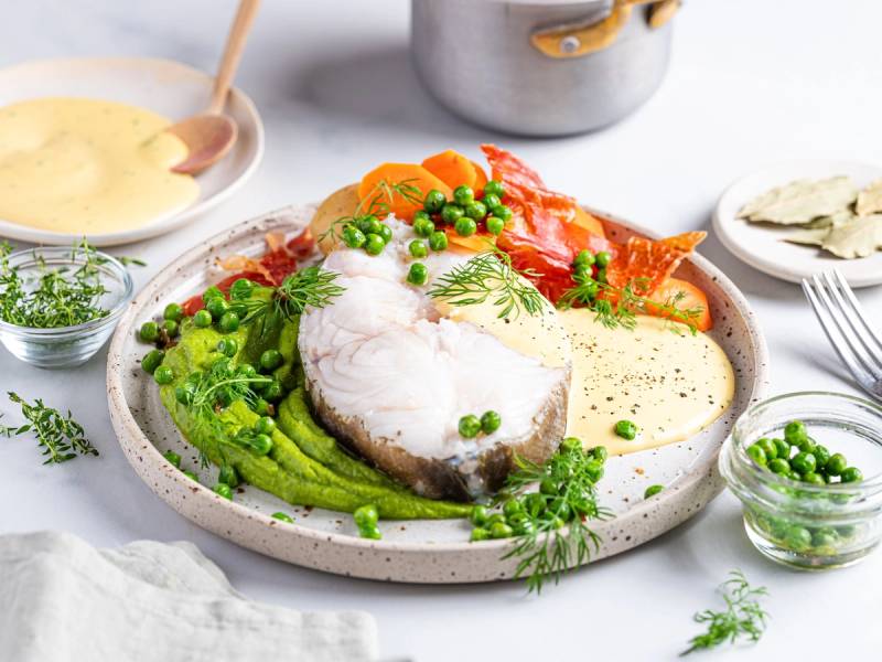 Skrei med spekeskinke og hollandaise-saus servert med grønnsaker og urter på en tallerken.