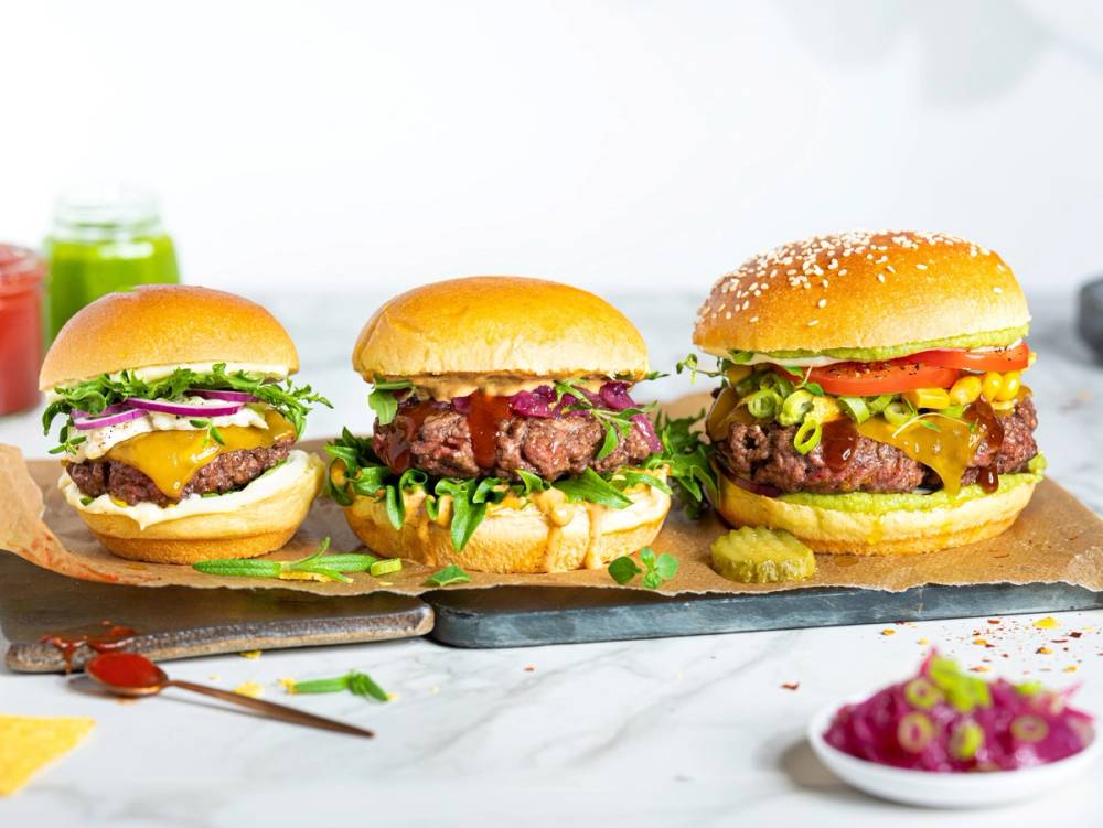 Tre sommerburger med ulike fyll på et skjærebrett, omgitt av små tilbehør og grønnsaker.