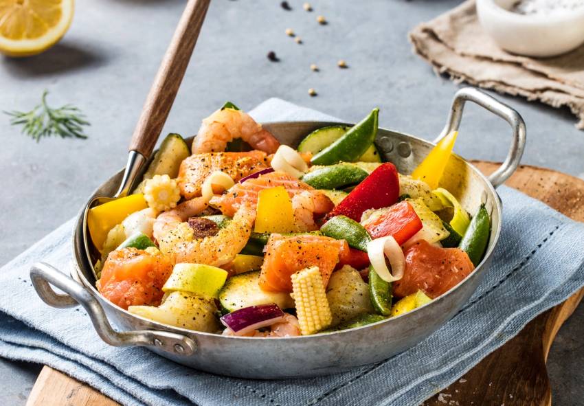 En wok med fisk og grønnsaker som paprika, squash og gulrot, servert i en metallpanne på et bord.