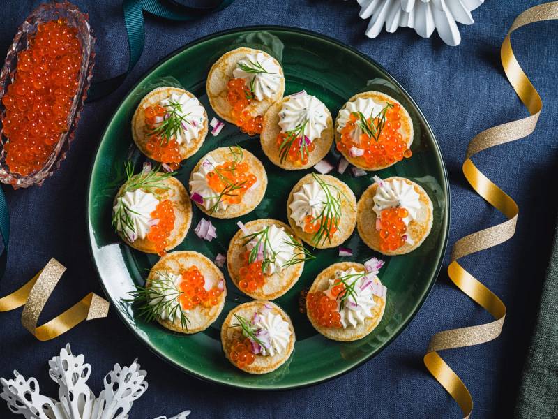 Små blinis med pisket rømme og røde kaviar på en grønn tallerken, pyntet med urter og dekorative elementer rundt.