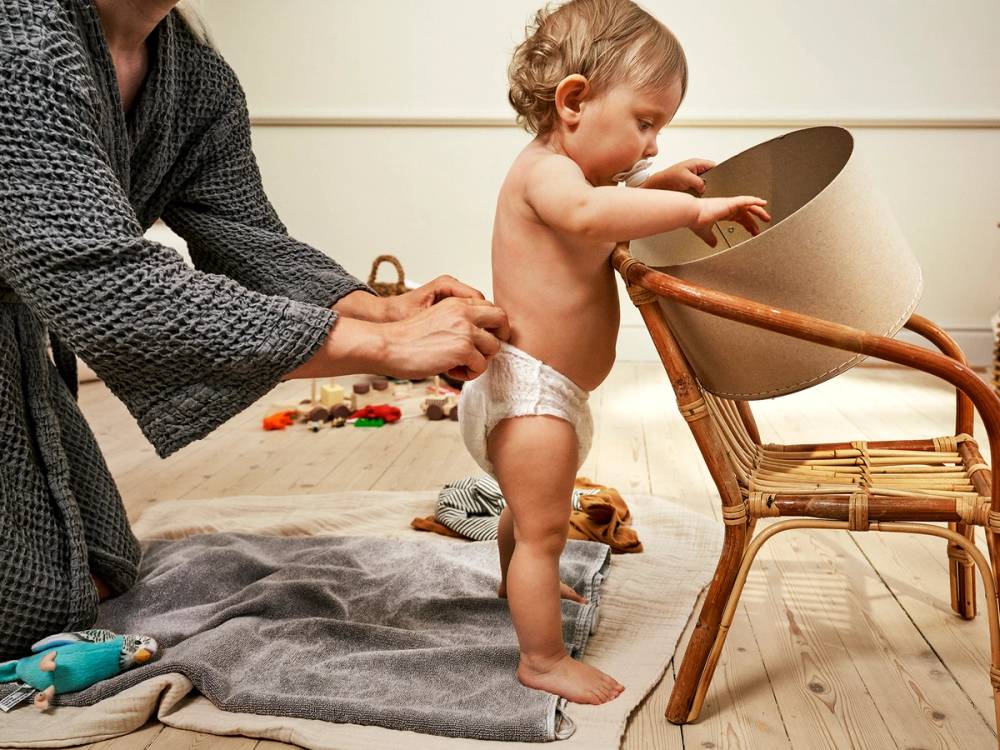 En liten baby i bleie som står oppreist og holder i en stol, mens en voksen hjelper eller passer på, på et rom med leker på gulvet.