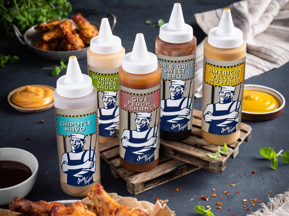 Fire flasker med ulike sauser på en treplate, omgitt av pizza og små skåler med saus, på et mørkt bord.