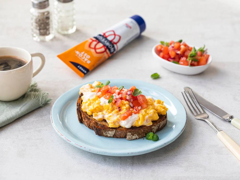 En skive grovt brød med smøreost og eggerøre, servert på en tallerken med en skål tomatsalat ved siden av.