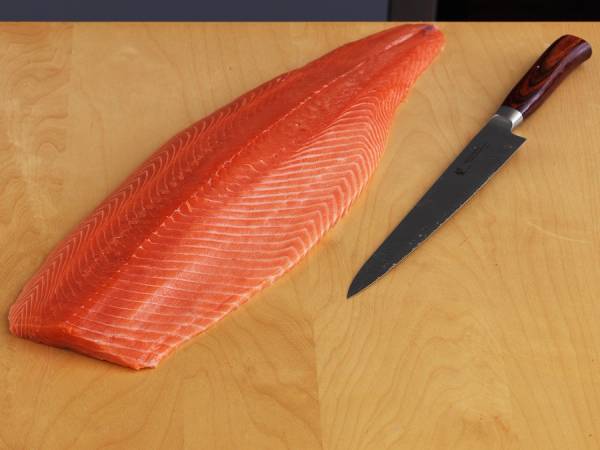 Fersk laksefilet ligger på et lyst trebord med en kjøkkenkniv ved siden av.