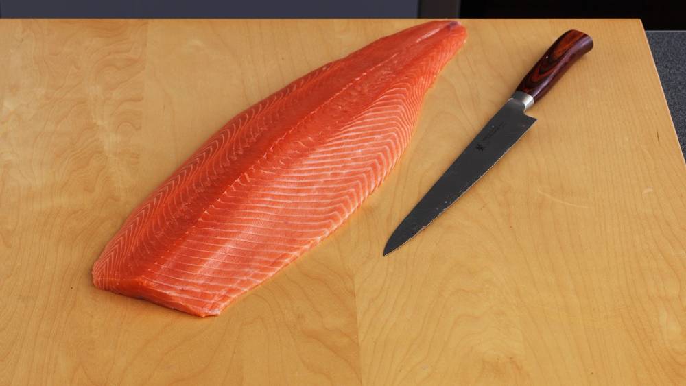 Fersk laksefilet ligger på et lyst trebord med en kjøkkenkniv ved siden av.