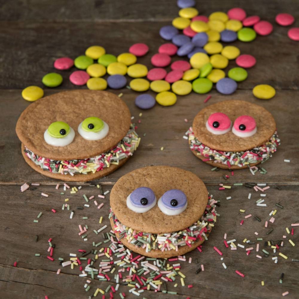 Tre pepperkaker med fargerike godteriøyne og sprinkles, omgitt av små fargede sjokoladeknapper på en treplate