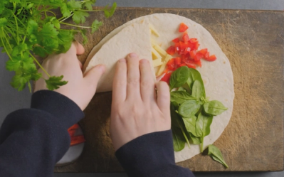 En person legger fylte ingredienser på en tortilla før rulling, med friske urter og grønnsaker rundt.