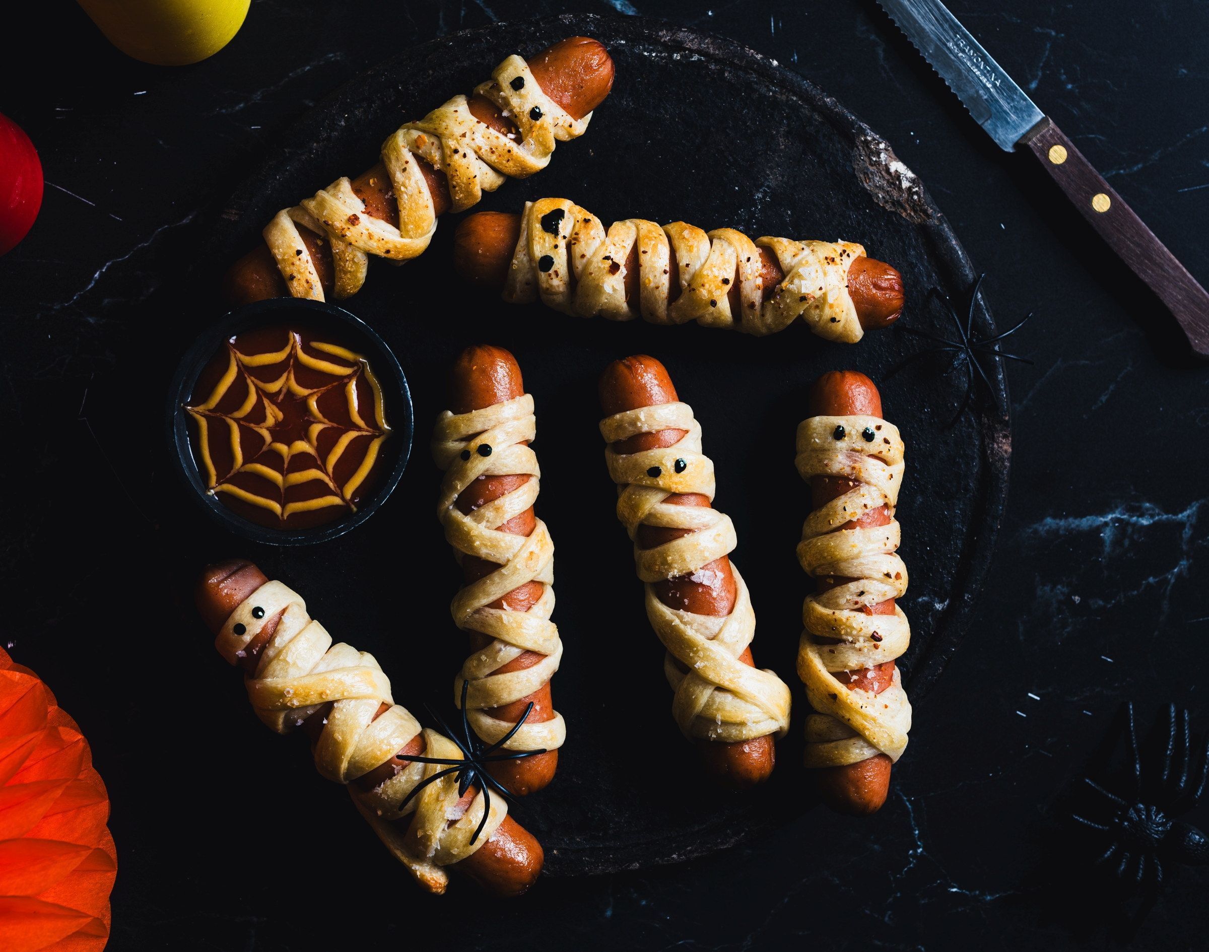Pølsepynt med spindelvev og små spøkelser laget av mat, satt på et svart fat til Halloween-feiring.