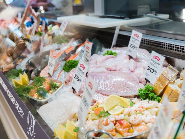 Fersk fisk og sjømat utstilt på en fiskedisk med priser og garnityr som sitron og grønt, i en dagligvarebutikk.