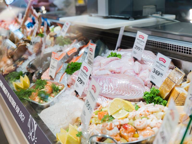 Fersk fisk og sjømat utstilt på en fiskedisk med priser og garnityr som sitron og grønt, i en dagligvarebutikk.