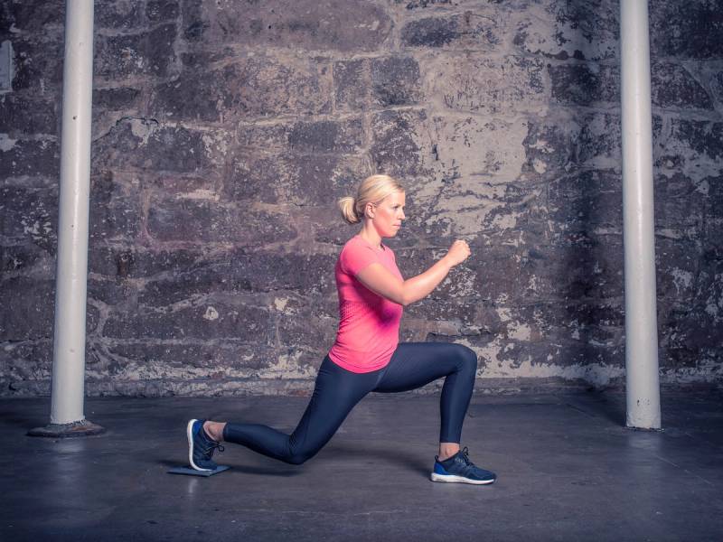 En person i rosa treningtøy gjør en utfalløvelse med sklimatte i et treningsrom, med en murvegg i bakgrunnen.