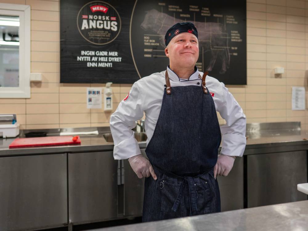 En mann i arbeidsantrekk står bak en kjøkkenbenk i en butikk, med en meny og informasjon på veggen bak ham.