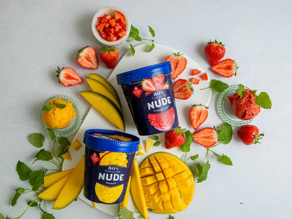 To isbokser med HennigOlsen NUDE iskrem omgitt av jordbær, bananer, sitron og mynte på en hvit bakgrunn.