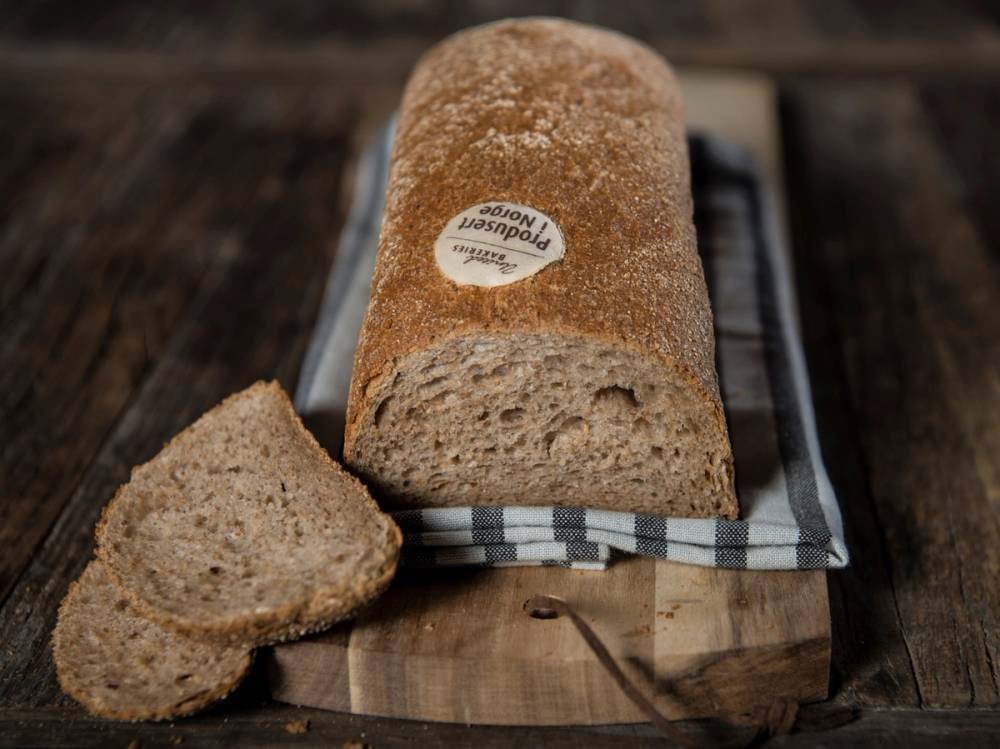 Grovt bondebrød med en skive tatt ut, plassert på et skjærebrett med et kjøkkenhåndkle under, på et treverk.